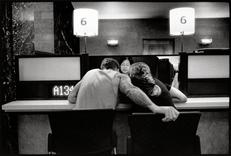 A man and a woman sit at a service counter, leaning toward a bank teller while the man rests his arm on the back of the woman's chair.