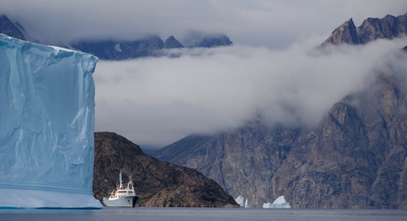 A white expedition ship sails on calm water past a massive blue iceberg, set against a backdrop of jagged, cloud-covered mountains.