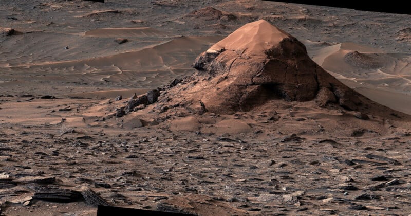 A large, conical rock formation sits in a vast, rocky Martian landscape covered in scattered stones and wind-swept sand dunes.