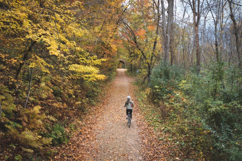 A person wearing a helmet rides a bicycle away from the camera along a leaf-covered trail through an autumn forest toward a distant stone tunnel.