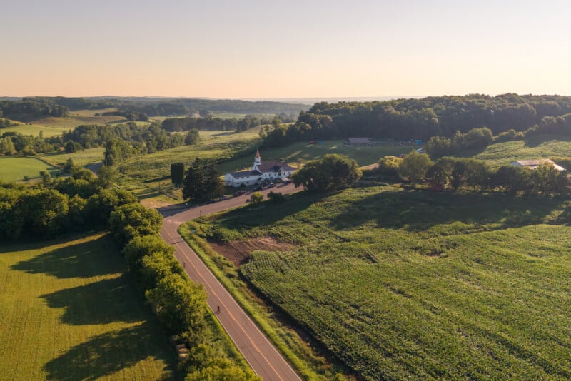 An aerial view shows a rural landscape with a church, a road winding past cornfields, and a wooded area under a clear sky.