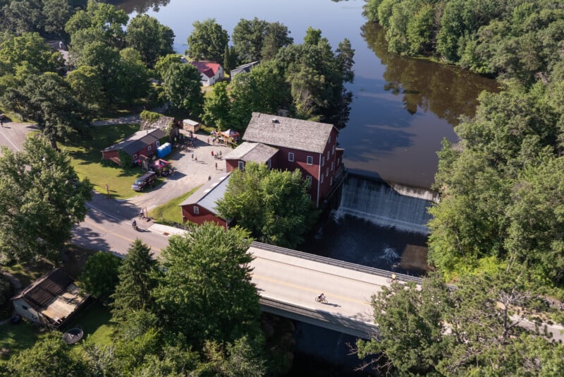 An aerial view shows a red mill building next to a waterfall and a bridge where cyclists are riding over a river.