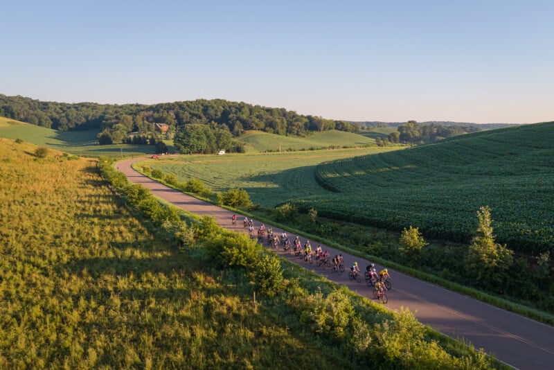 A large group of cyclists rides in a line along a winding paved road through a rural landscape of rolling green hills and fields.