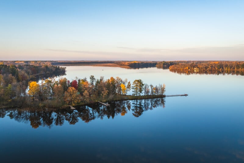 An aerial view of a forested point of land extending into a calm lake during autumn, with colorful trees reflecting in the water.