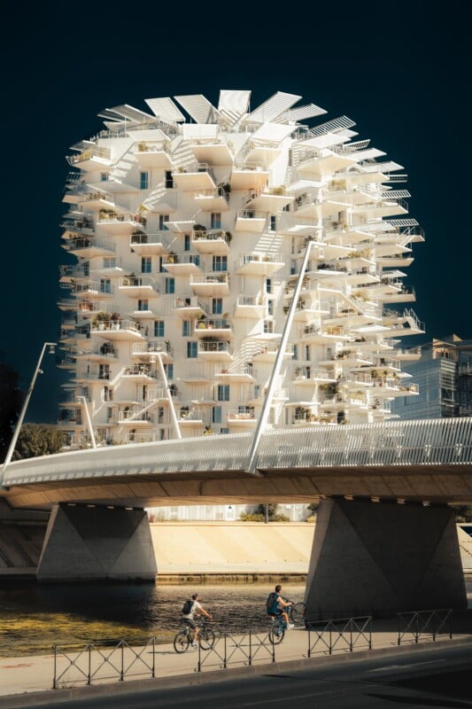 Two people ride bicycles on a path beside a river under a bridge, with a tall, white, modern apartment building featuring many cantilevered balconies in the background.