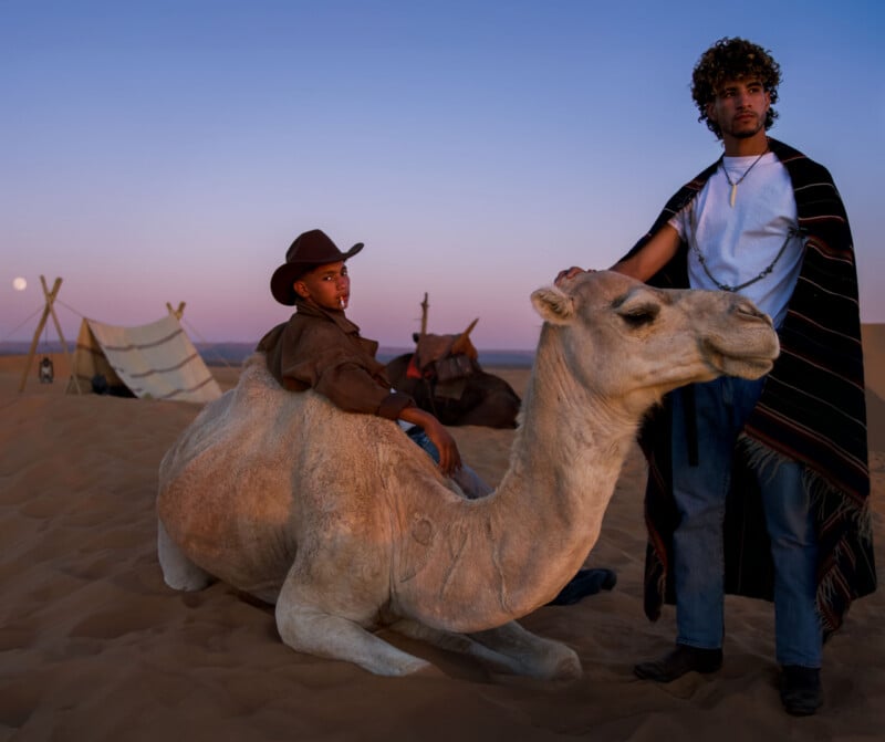 A man with curly hair stands beside a seated camel, while a person wearing a cowboy hat sits on the camel's back in a desert at dusk.