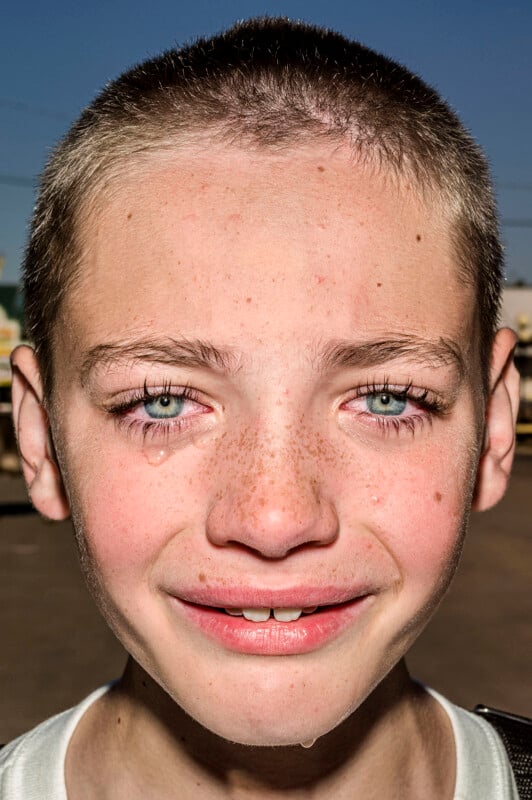 A close-up portrait of a young person with short hair, freckles, and blue eyes, with a single tear running down their cheek.