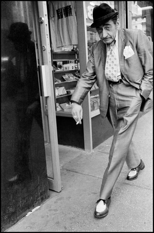 An older man in a suit, hat, and two-toned shoes walks past a storefront while holding a cigarette.