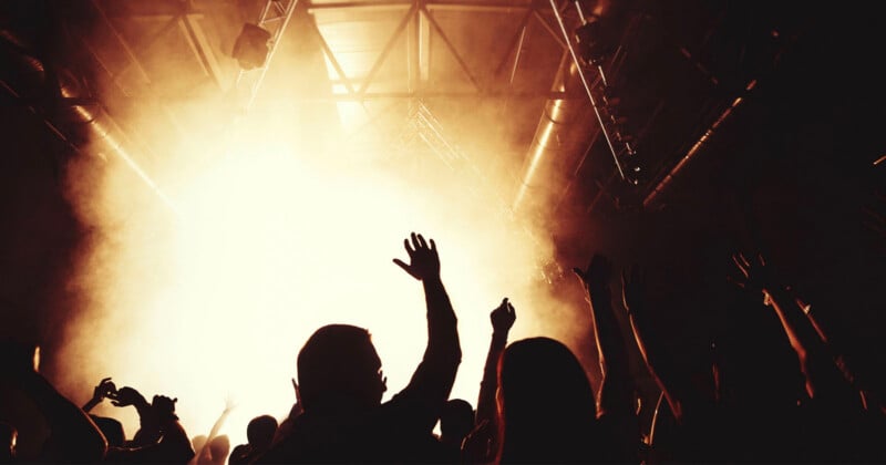 Silhouetted concertgoers raise their hands toward a bright, hazy stage light in a dark venue.