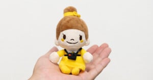 A small, plush doll with brown hair in a bun and a yellow dress, wearing a miniature black camera around its neck, sits in an open palm.