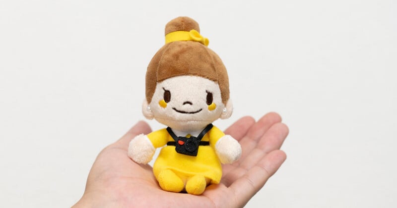 A small, plush doll with brown hair in a bun and a yellow dress, wearing a miniature black camera around its neck, sits in an open palm.