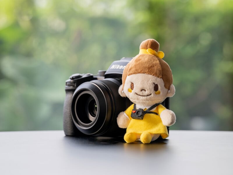 A plush doll with brown hair in a bun and a yellow dress sits next to a black Nikon camera on a smooth surface against a blurred green background.