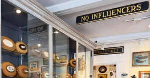 A shop interior features a sign reading "NO INFLUENCERS" hanging above glass display cases filled with round, woven Nantucket-style baskets.