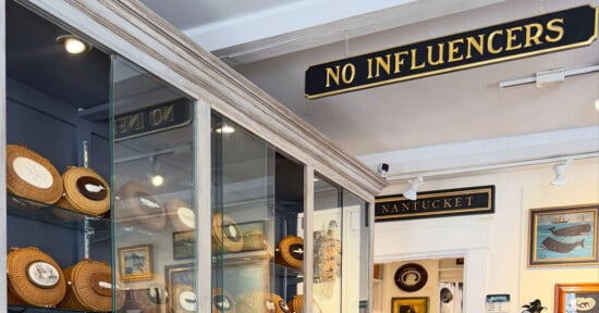 A shop interior features a sign reading "NO INFLUENCERS" hanging above glass display cases filled with round, woven Nantucket-style baskets.