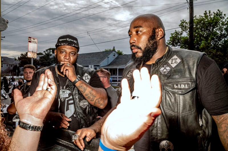 Two men wearing leather motorcycle club vests with "1%" patches stand outdoors, while a person's hands are held up in the foreground.