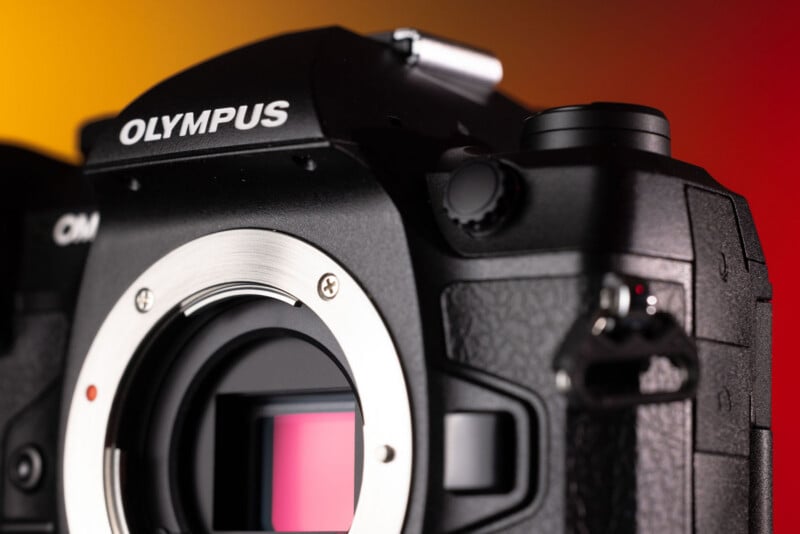 A close-up view of a black Olympus OM-D camera body, focusing on the lens mount and the sensor against a yellow and red background.