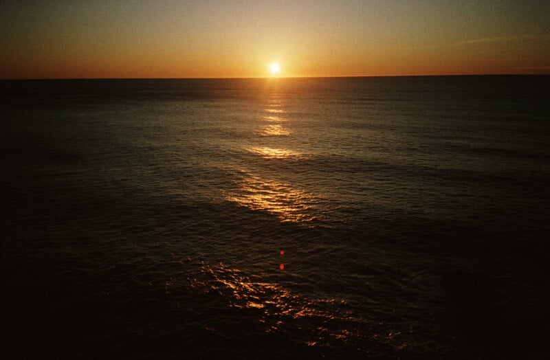 The sun sets over a dark, rippling ocean, casting a bright golden reflection across the water's surface.