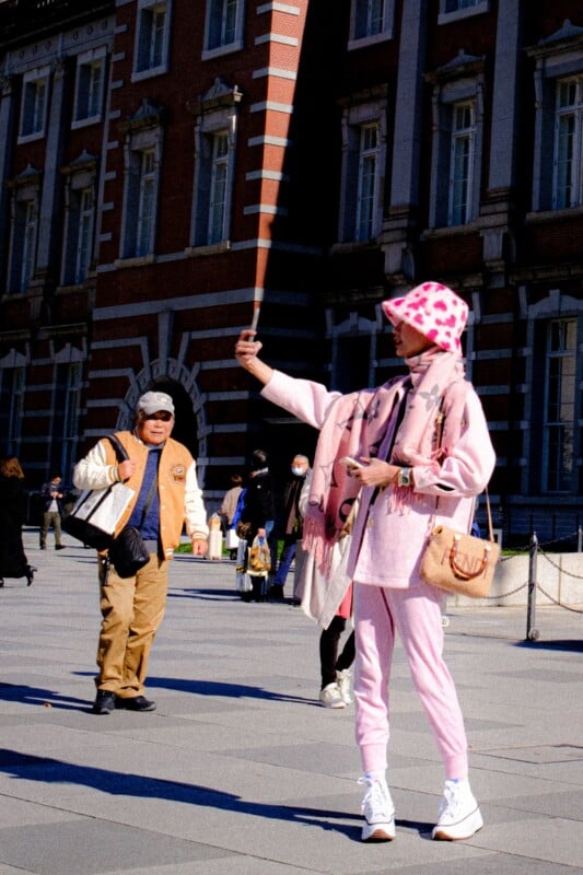 A person in a pink patterned bucket hat and matching outfit takes a selfie in front of a brick building, while a man in a tan jacket walks nearby.