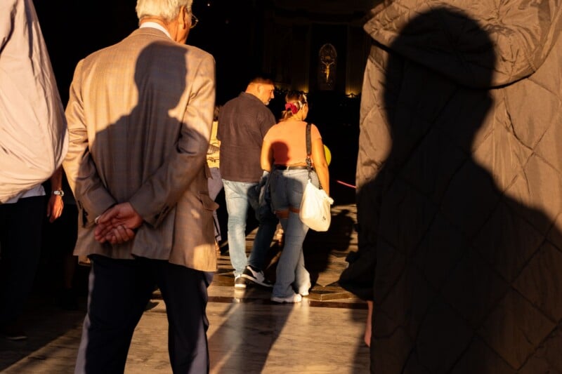 People walk through a sunlit, shadowed interior space toward a distant, illuminated crucifix on a wall.