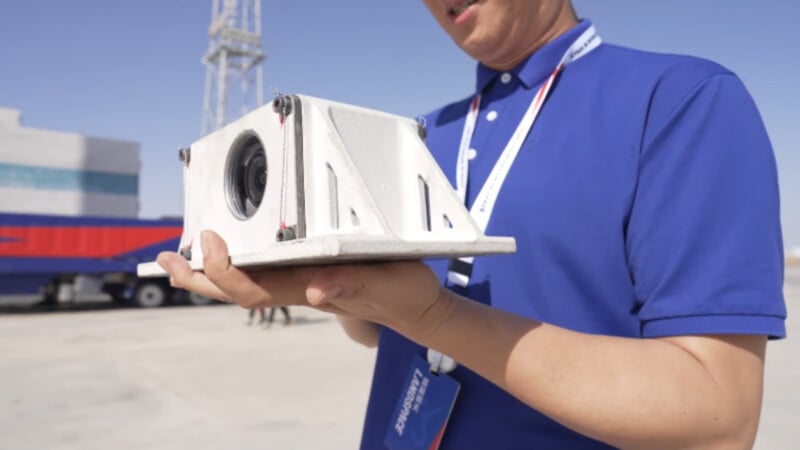A person in a blue polo shirt holds a white, wedge-shaped camera housing with a lens visible on the front face.
