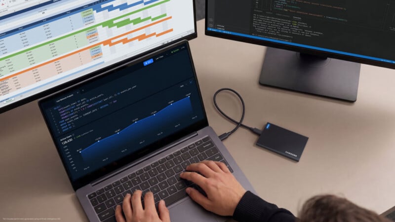 A person types on a laptop connected to an external hard drive, with two monitors displaying project management charts and code in the background.