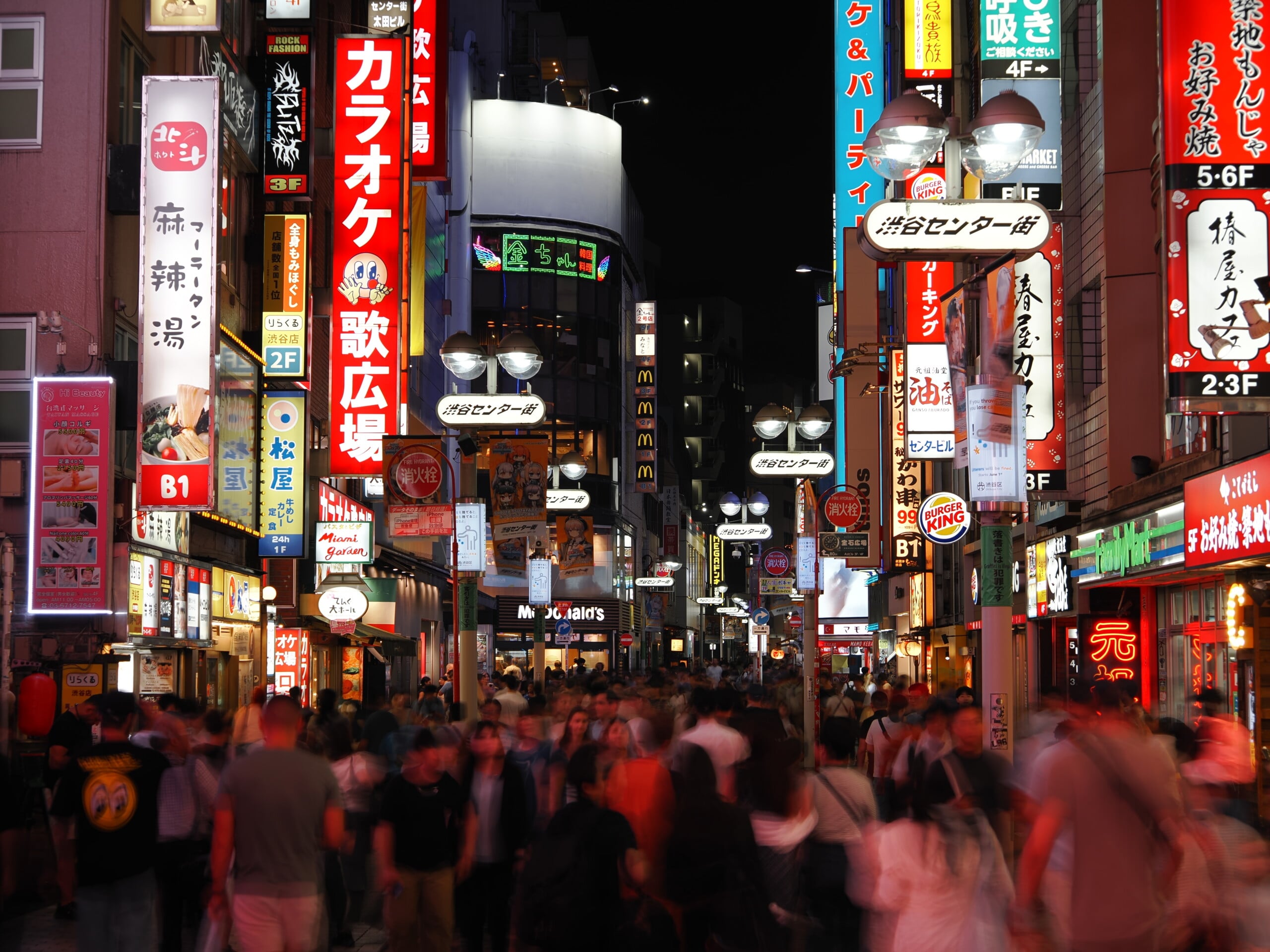 A crowded street in Tokyo at night, filled with bright, multi-colored neon signs and advertisements in Japanese.