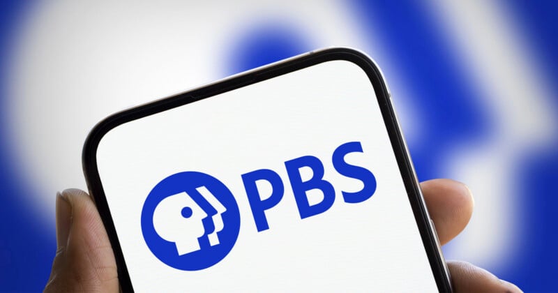 A hand holds a smartphone displaying the blue PBS logo on a white screen against a blurred blue and white background.