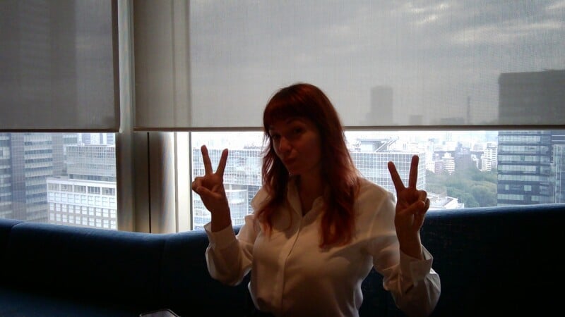 A woman with reddish-brown hair and bangs wearing a white button-down shirt makes peace signs with both hands while sitting in front of a window overlooking a city.