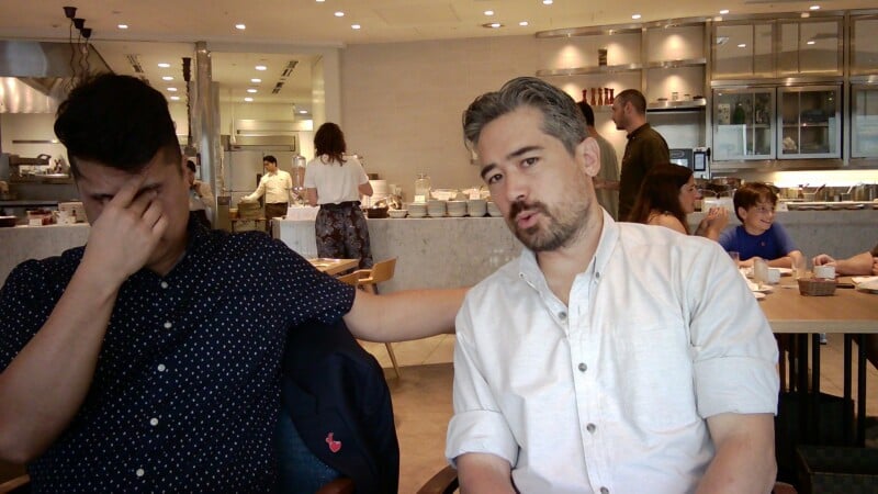 A man in a white button-down shirt sits at a restaurant table with his arm around a man in a dark patterned shirt who is covering his face.