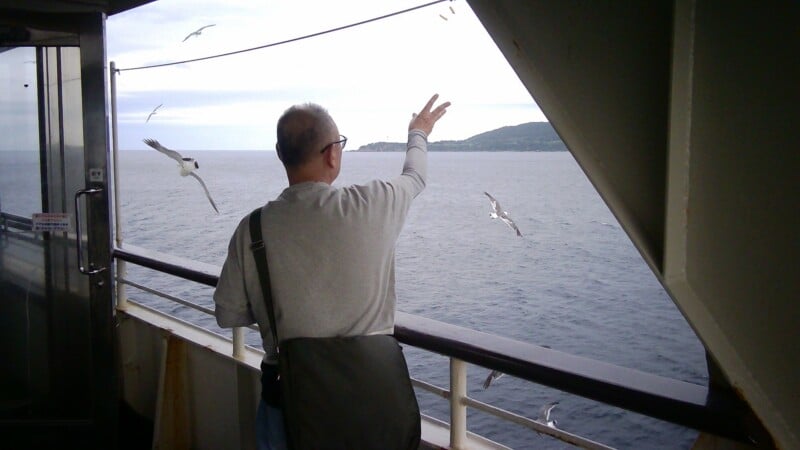 A man in a grey long-sleeved shirt and a shoulder bag stands on a ferry deck, reaching out to feed seagulls flying over the ocean.