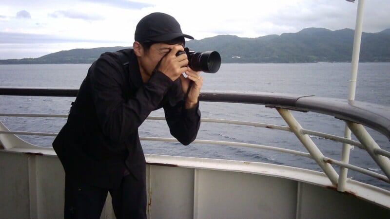 A person wearing a black shirt and cap stands on a boat deck, holding a camera up to their eye to take a photograph of the sea and distant mountains.