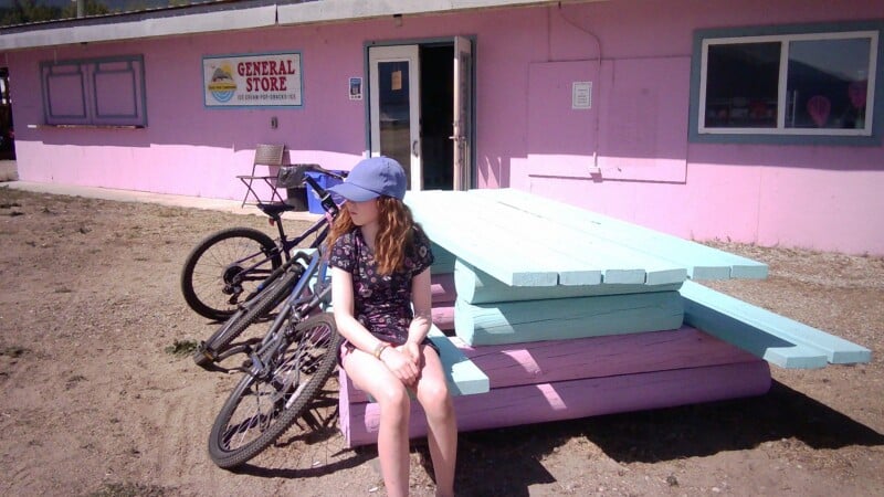 A girl wearing a blue baseball cap and a floral dress sits on a light blue and pink log bench in front of a pink general store.