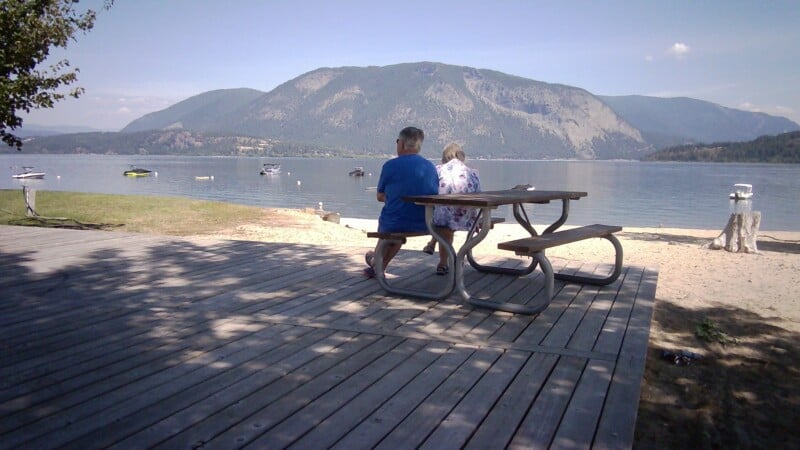 An older couple sits at a wooden picnic table on a deck overlooking a lake with mountains in the background.