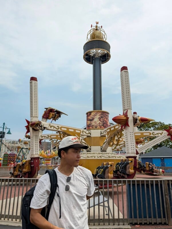 A young man wearing a white t-shirt and a baseball cap stands in front of a large, mechanical amusement park ride.