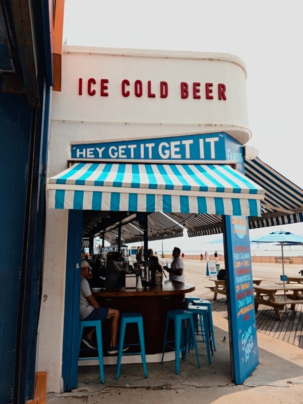 A beachside bar with a blue and white striped awning, red "ICE COLD BEER" signage, and patrons sitting on blue stools at a wooden counter.
