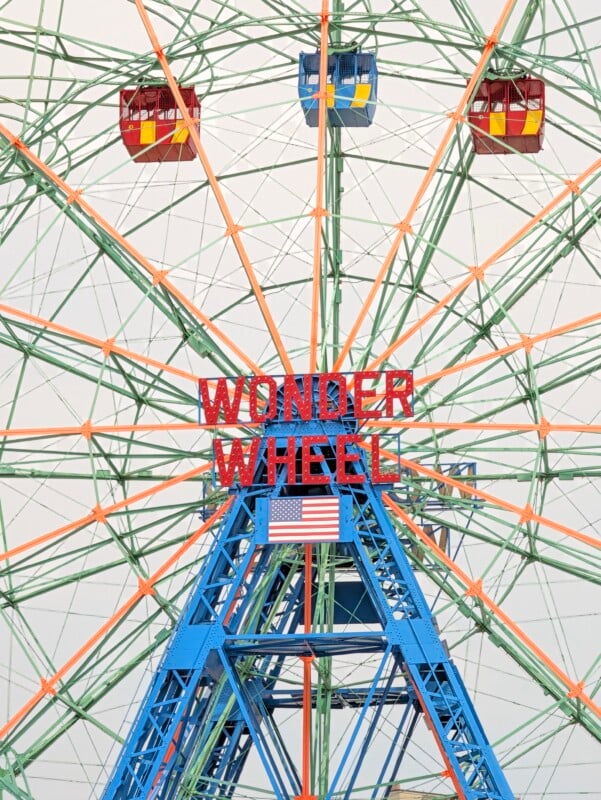 The Wonder Wheel ferris wheel in Coney Island features red and blue passenger cars, a central sign reading "WONDER WHEEL," and an American flag.