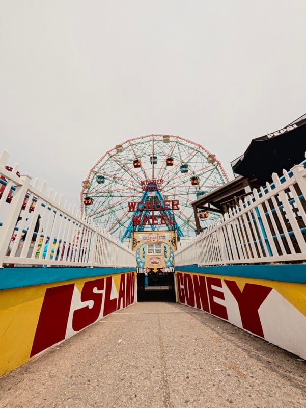 A path leads toward the Wonder Wheel at Coney Island, framed by white picket fences and a yellow wall with red lettering reading "ISLAND CONEY.