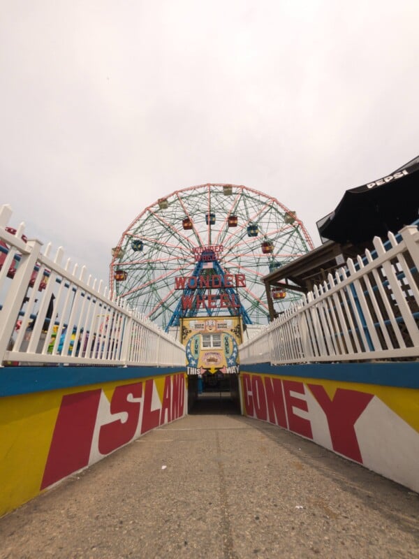 A paved path leads toward the Wonder Wheel at Coney Island, framed by white picket fences and yellow walls with red lettering reading "ISLAND" and "CONEY.