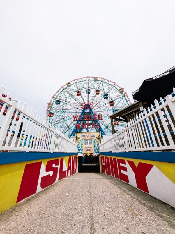 A path leads toward the Wonder Wheel at Coney Island, framed by white fences and walls painted with the words "ISLAND" and "CONEY.