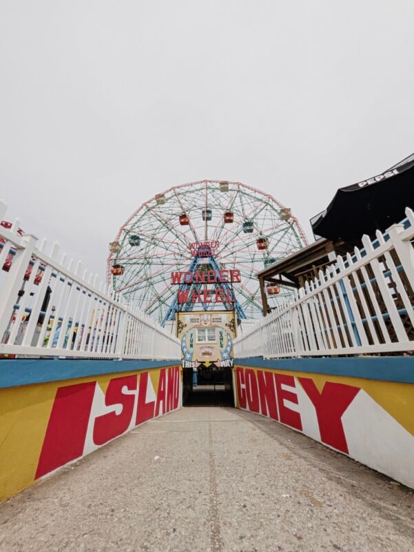 A path leads toward the Wonder Wheel at Coney Island, framed by white picket fences and a yellow wall with red lettering reading "ISLAND CONEY.