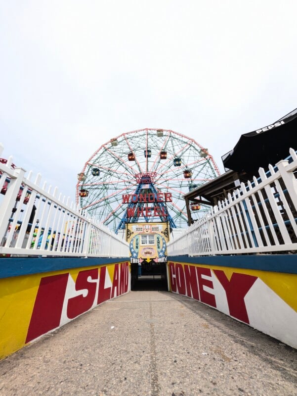 A path leads through a tunnel entrance with "ISLAND" and "CONEY" painted on the walls, toward the Wonder Wheel at an amusement park.