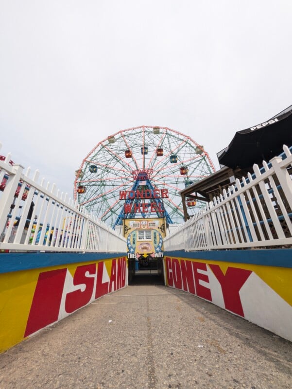 A path leads toward the Wonder Wheel at Coney Island, framed by white picket fences and walls painted with the words "ISLAND" and "CONEY.