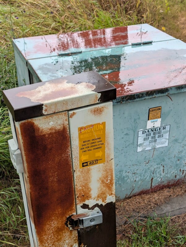 Two weathered, metal utility boxes sit in a grassy area, with the smaller foreground box featuring a yellow "Caution: Buried Cable" sign and the larger background box displaying "Danger: High Voltage" warning labels.