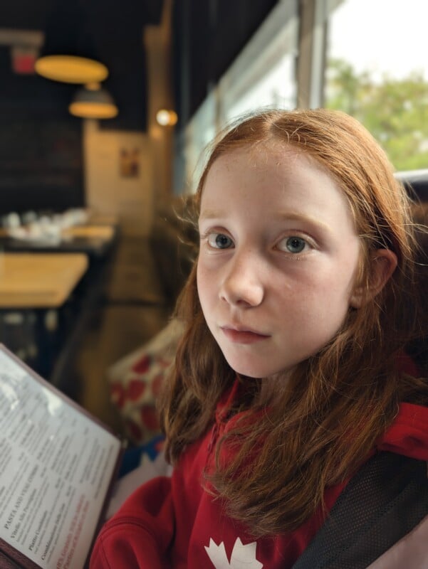 A young girl with long red hair and freckles looks toward the camera while holding a menu in a restaurant.