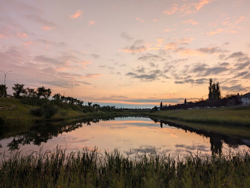 A calm pond reflects a pastel sunset sky with pink and orange clouds, framed by grassy banks and silhouetted trees.
