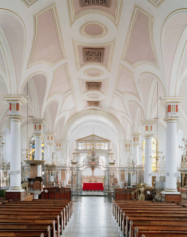 The interior of a church features rows of wooden pews leading toward a central altar covered with a red cloth, framed by ornate metal gates and white columns supporting a vaulted ceiling.