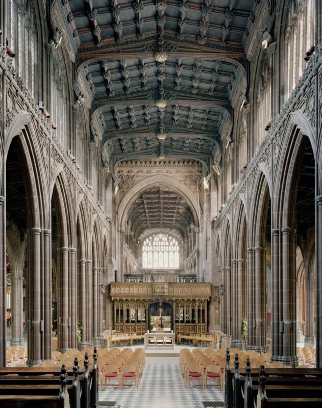 The interior of a Gothic-style church features a long nave with stone columns, a dark wooden vaulted ceiling, and a central aisle leading to an ornate wooden screen and altar.