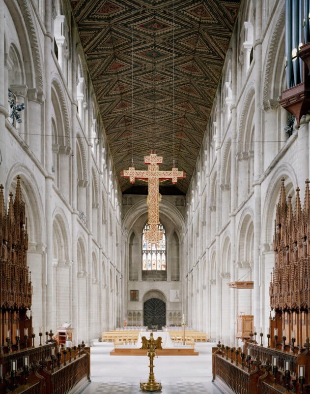 The interior of a long, stone cathedral nave features a high, patterned wooden ceiling, a large suspended cross, and rows of wooden pews leading to an altar.