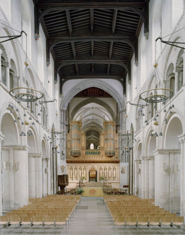 The interior of a stone church features rows of wooden chairs facing a central altar, a large pipe organ, and arched columns.