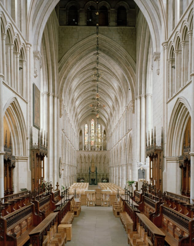 A symmetrical view down the center aisle of a stone cathedral, featuring rows of wooden choir stalls, a high vaulted ceiling, and stained glass windows at the far end.